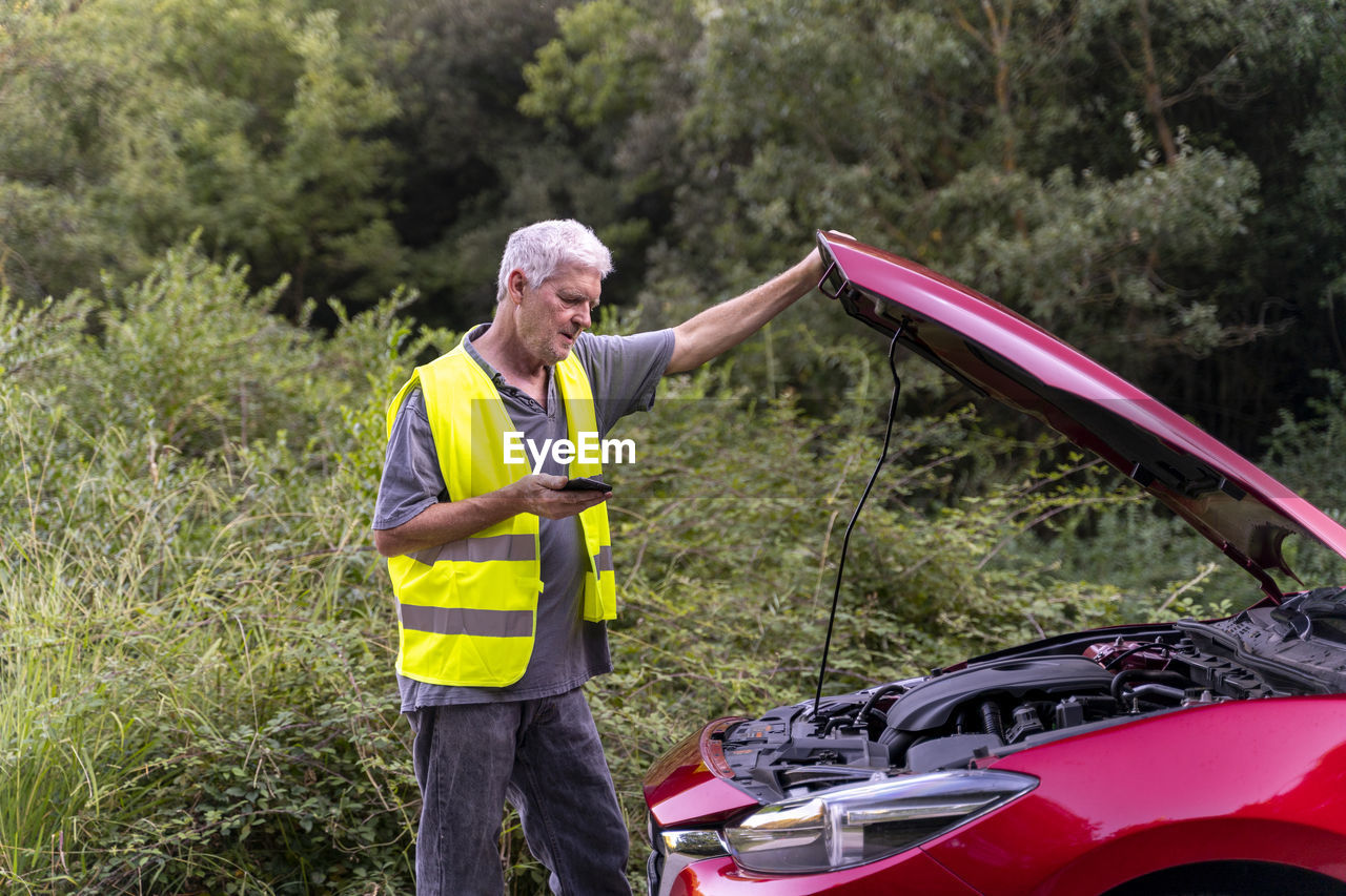 Senior man standing at his broken car wearing a safety vest and using his smartphone