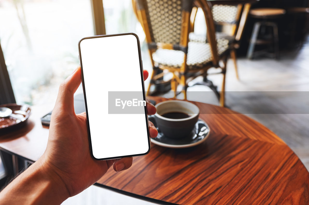 wireless technology, hand, cup, technology, coffee cup, coffee, communication, table, mug, food and drink, drink, portable information device, smartphone, one person, cafe, adult, mobile phone, business, indoors, holding, digital tablet, computer, refreshment, lifestyles, portability, restaurant, hot drink, internet, coffee shop, relaxation, day, crockery, tableware, activity, leisure activity, telephone, wood, computer network, women, screen, furniture