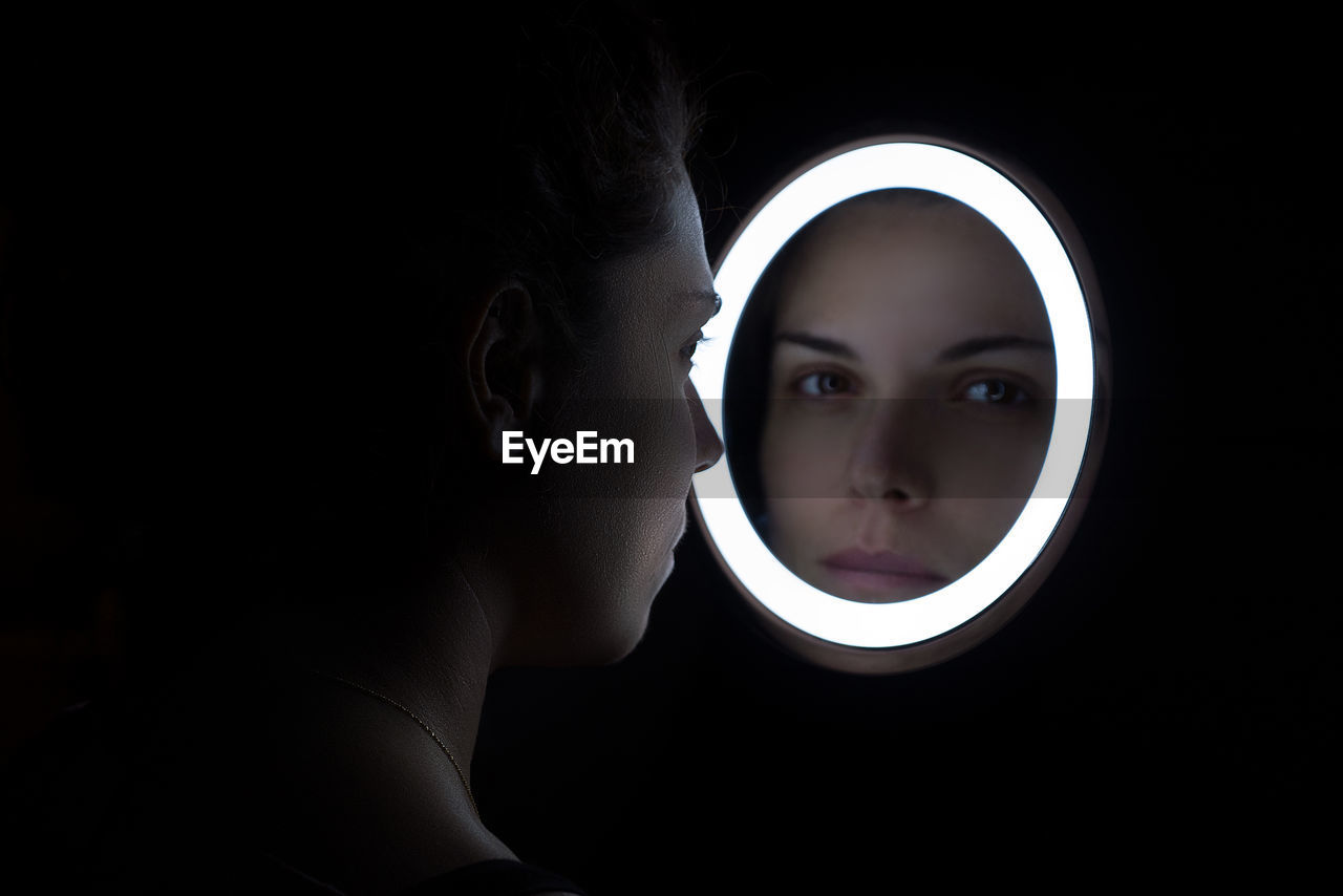 Close-up of young woman looking through mirror reflection over black background