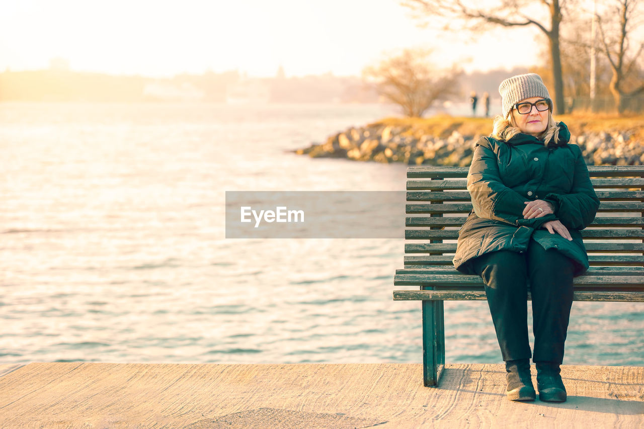 Full length of woman sitting on bench against sea in winter