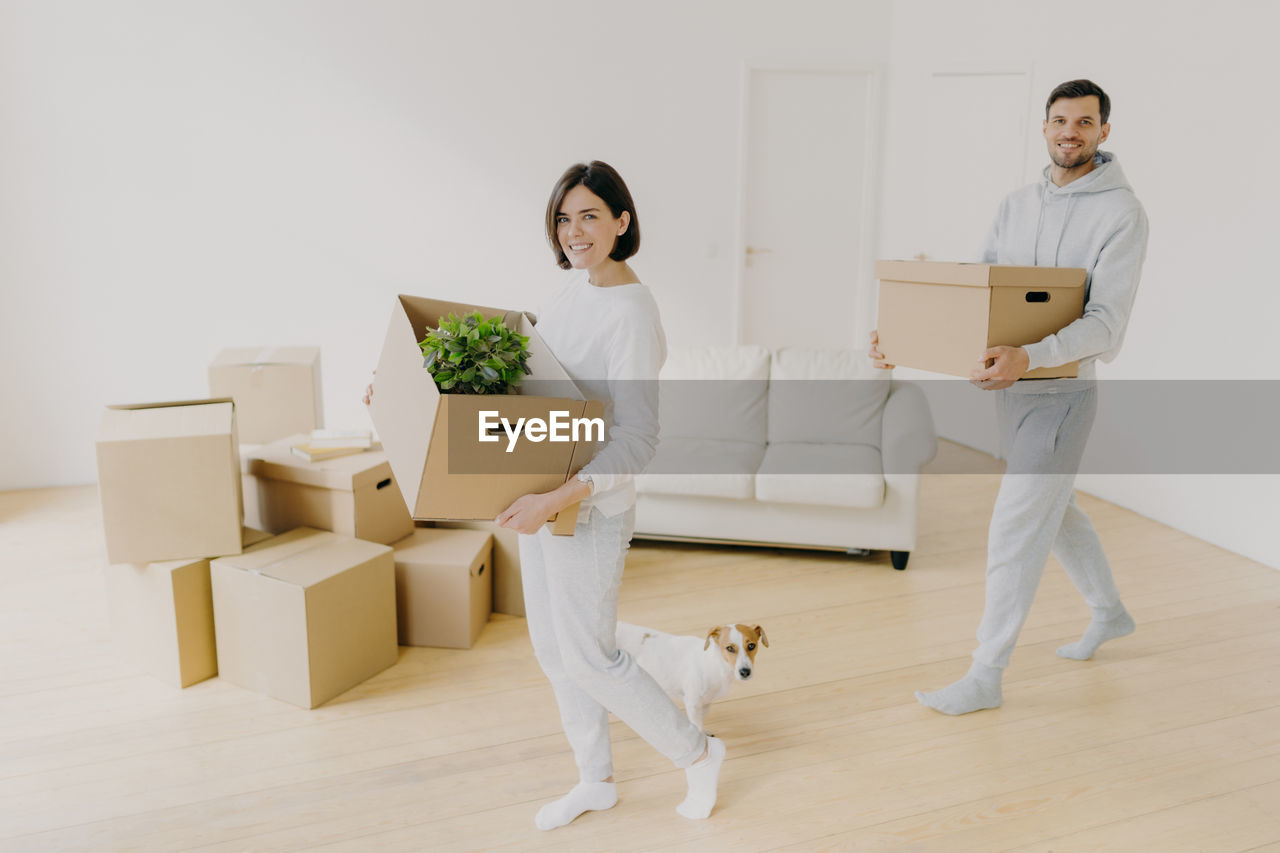 Smiling couple holding box standing on floor with dog