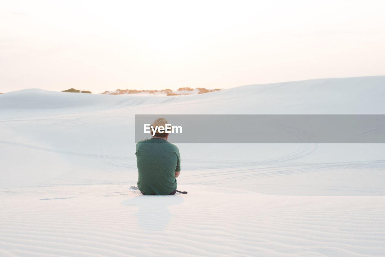 Rear view of man sitting on sand dune against clear sky