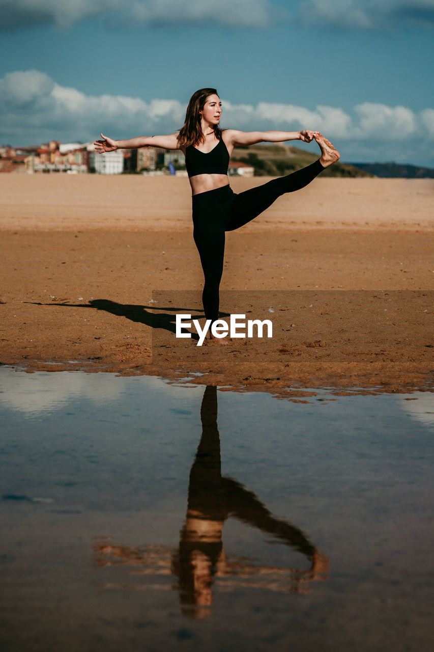 Side view of flexible female standing on sandy shore of river in extended hand to big toe pose and practicing yoga while balancing on leg with eyes closed