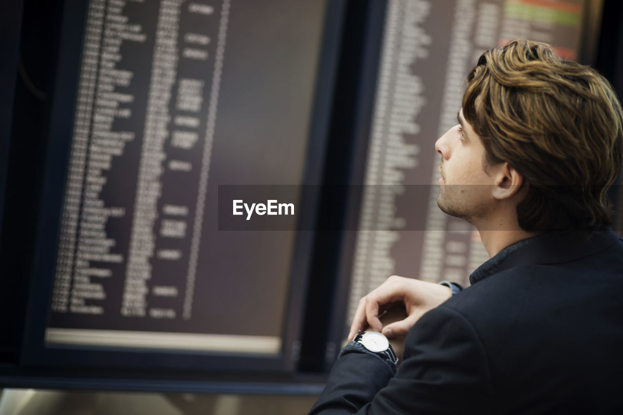 Businessman adjusting wrist watch while reading arrival departure board at airport