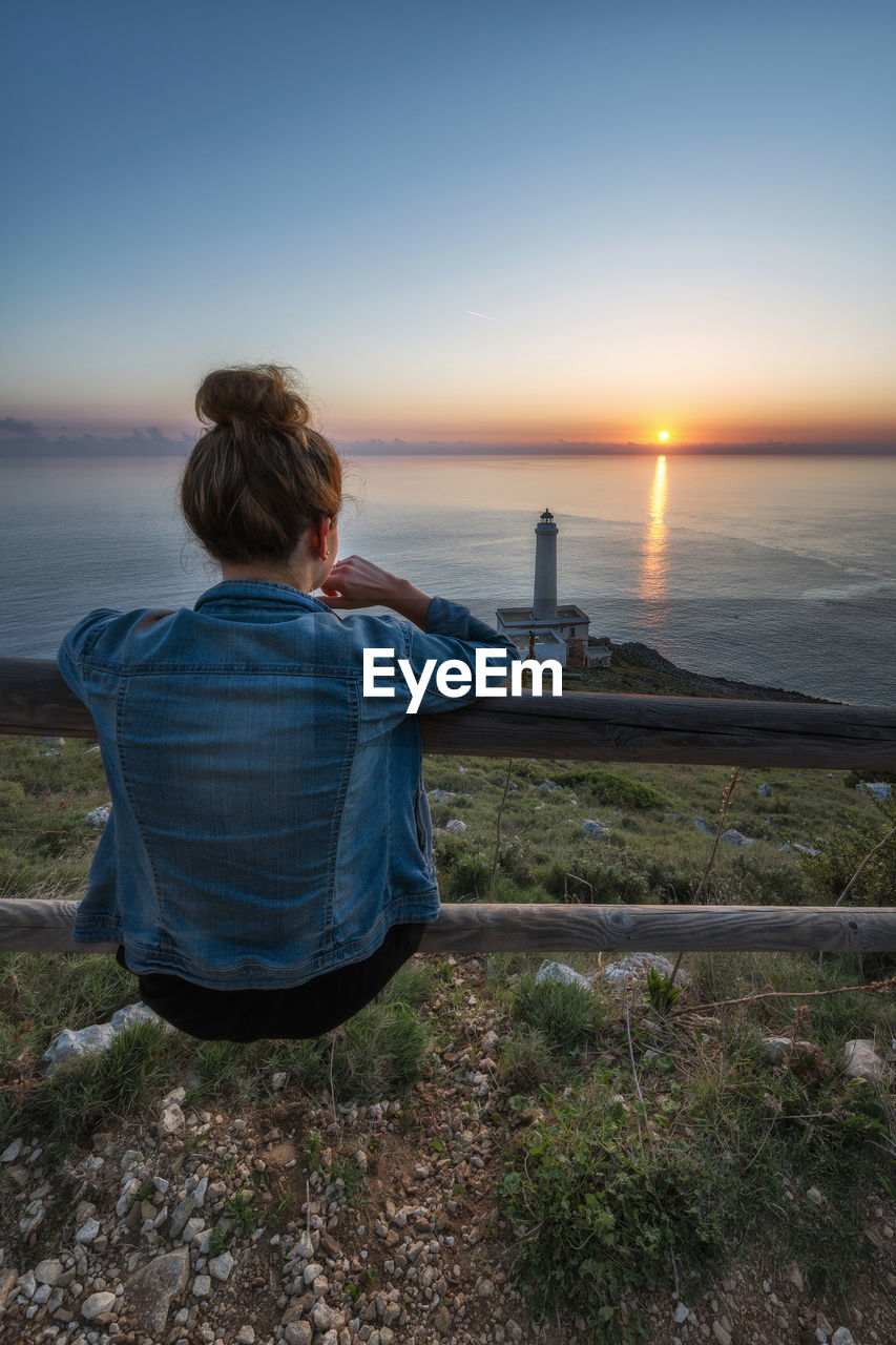 Rear view of woman sitting on railing while looking at sea against sky during sunset