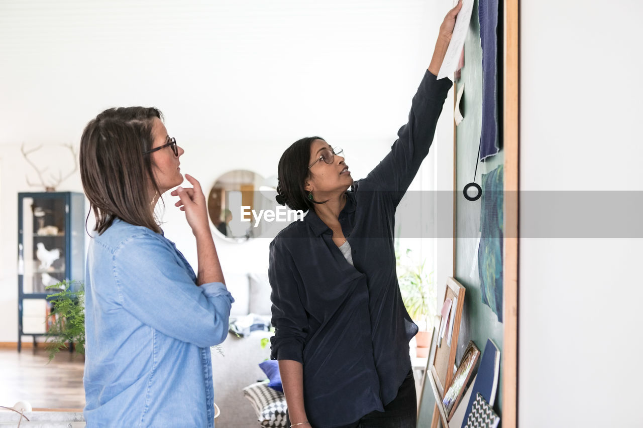 Female design professionals discussing over paintings on blackboard in home office