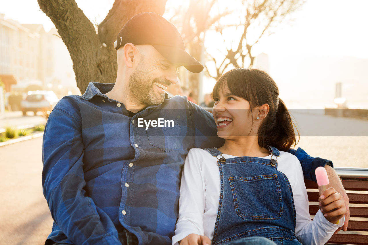 Cheerful father and daughter sitting on bench in city