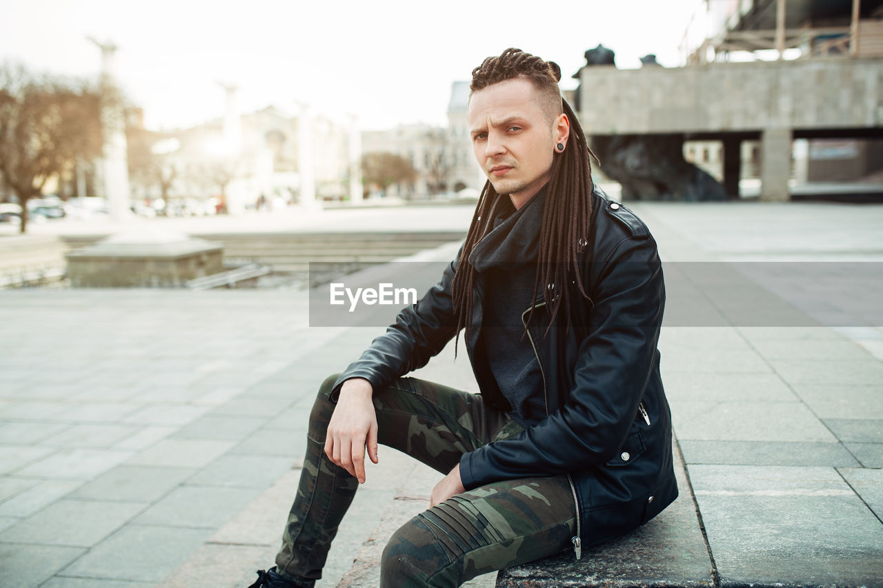 Portrait of hipster man sitting on footpath in city