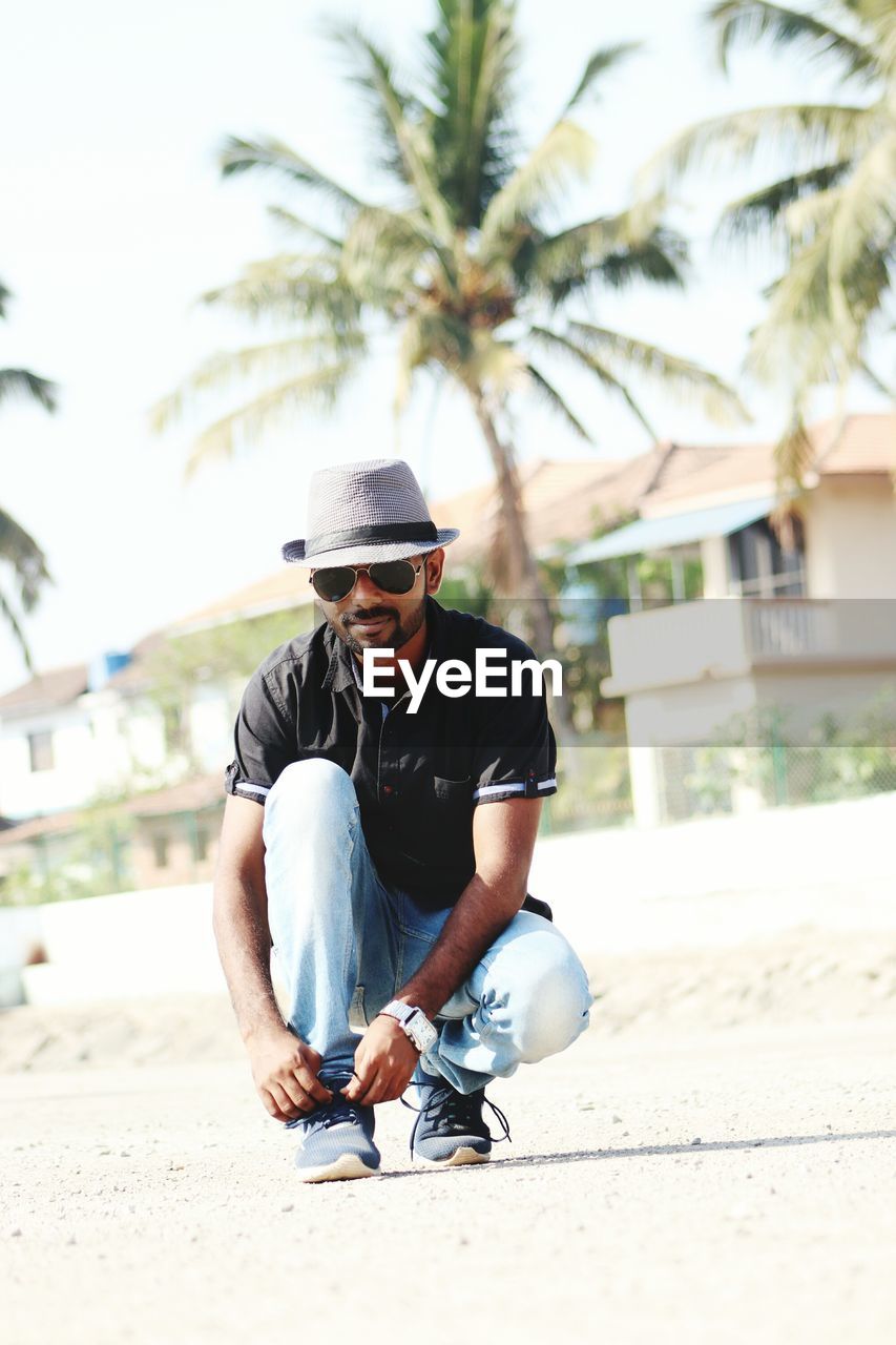 Portrait of man in sunglasses tying shoelace at beach against sky