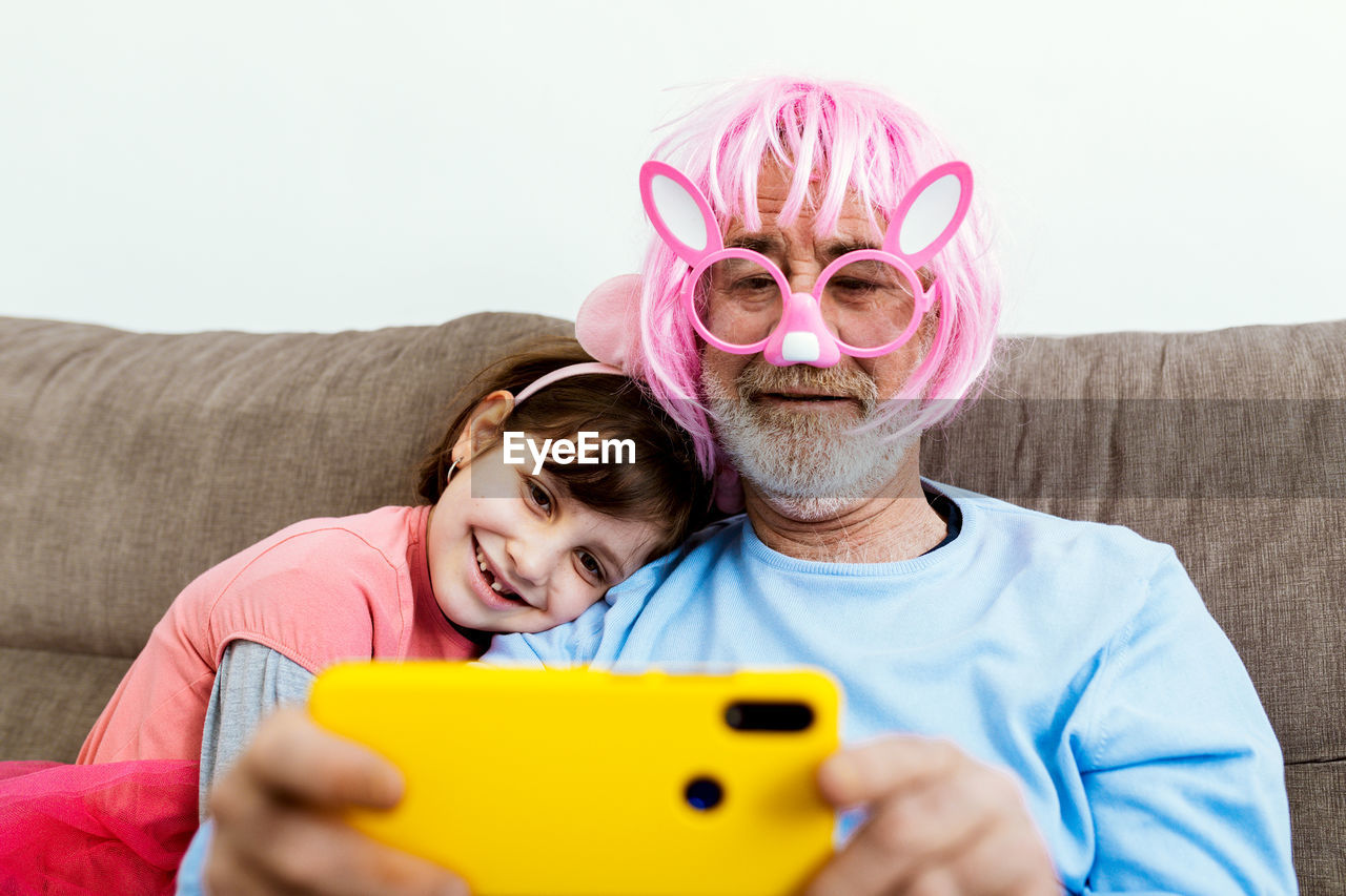 Happy child sitting on couch with grandfather in big pink plastic glasses and pink wig with false ears and making selfie on smartphone in room