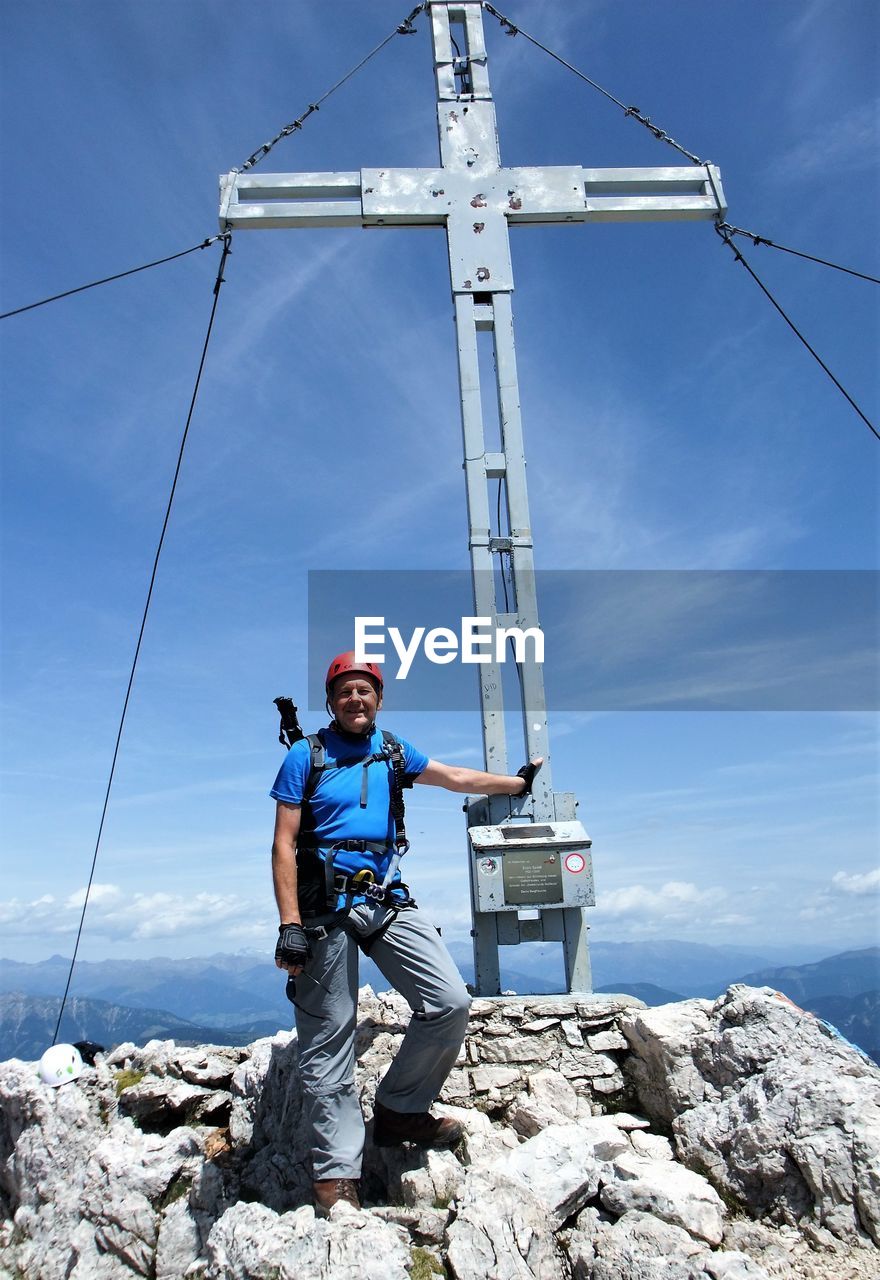 Male hiker standing by cross on cliff against blue sky