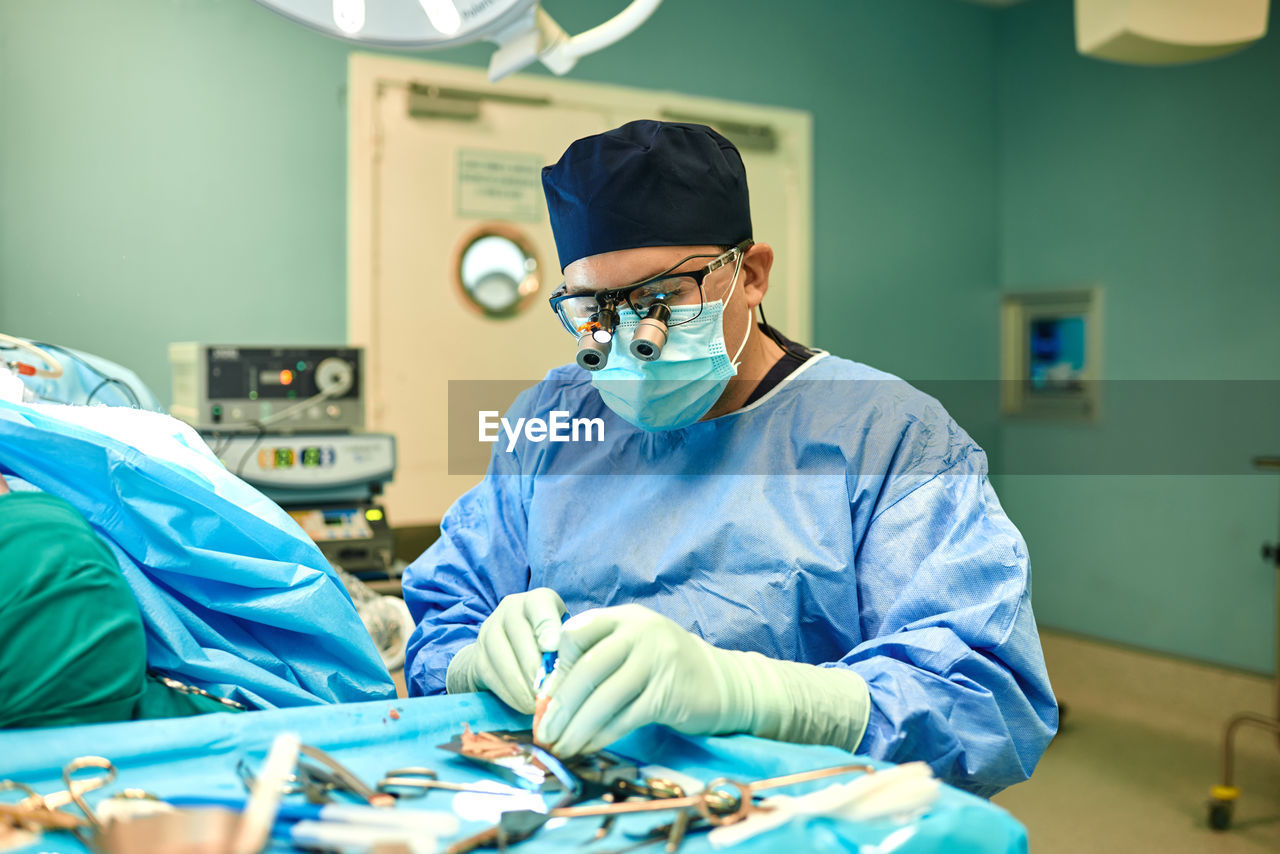 Anonymous focused male surgeon in blue gown mask and magnifying eyeglasses standing near sterile field with various medical tools during operation in modern clinic
