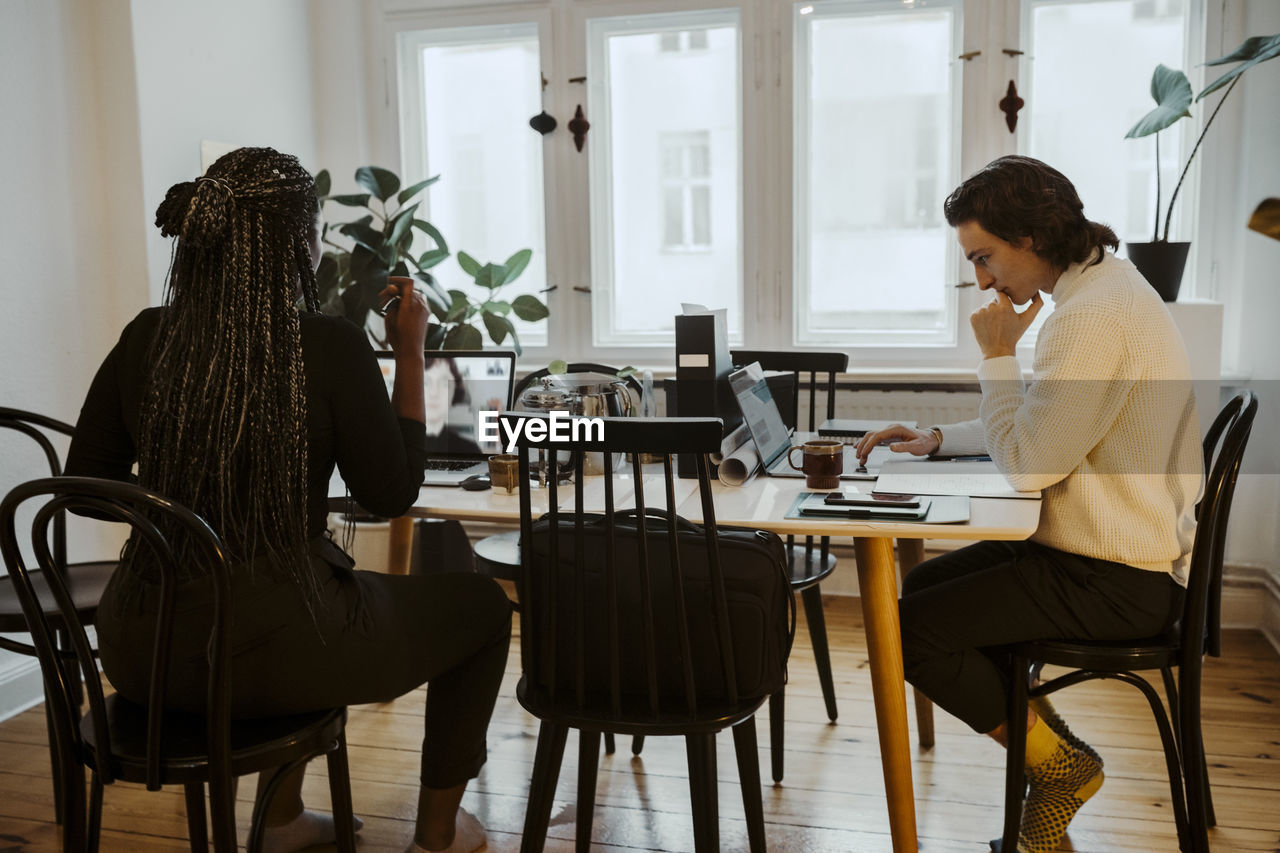 Businesswoman attending video call while male entrepreneur using laptop sitting at desk in home office