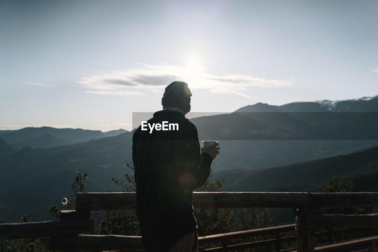 Rear view of man with cup of coffee looking at mountain range against sky