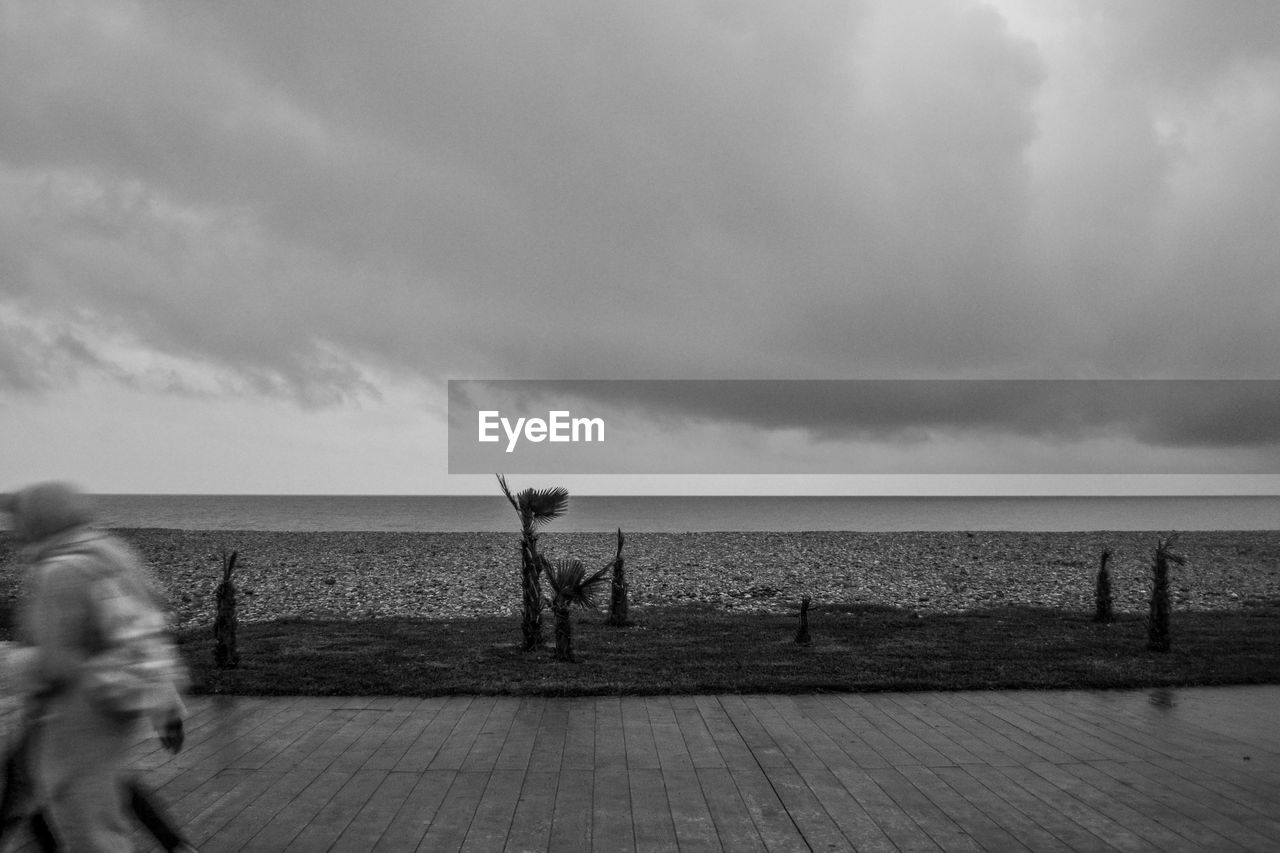 Blurred motion of woman walking on promenade by sea against cloudy sky