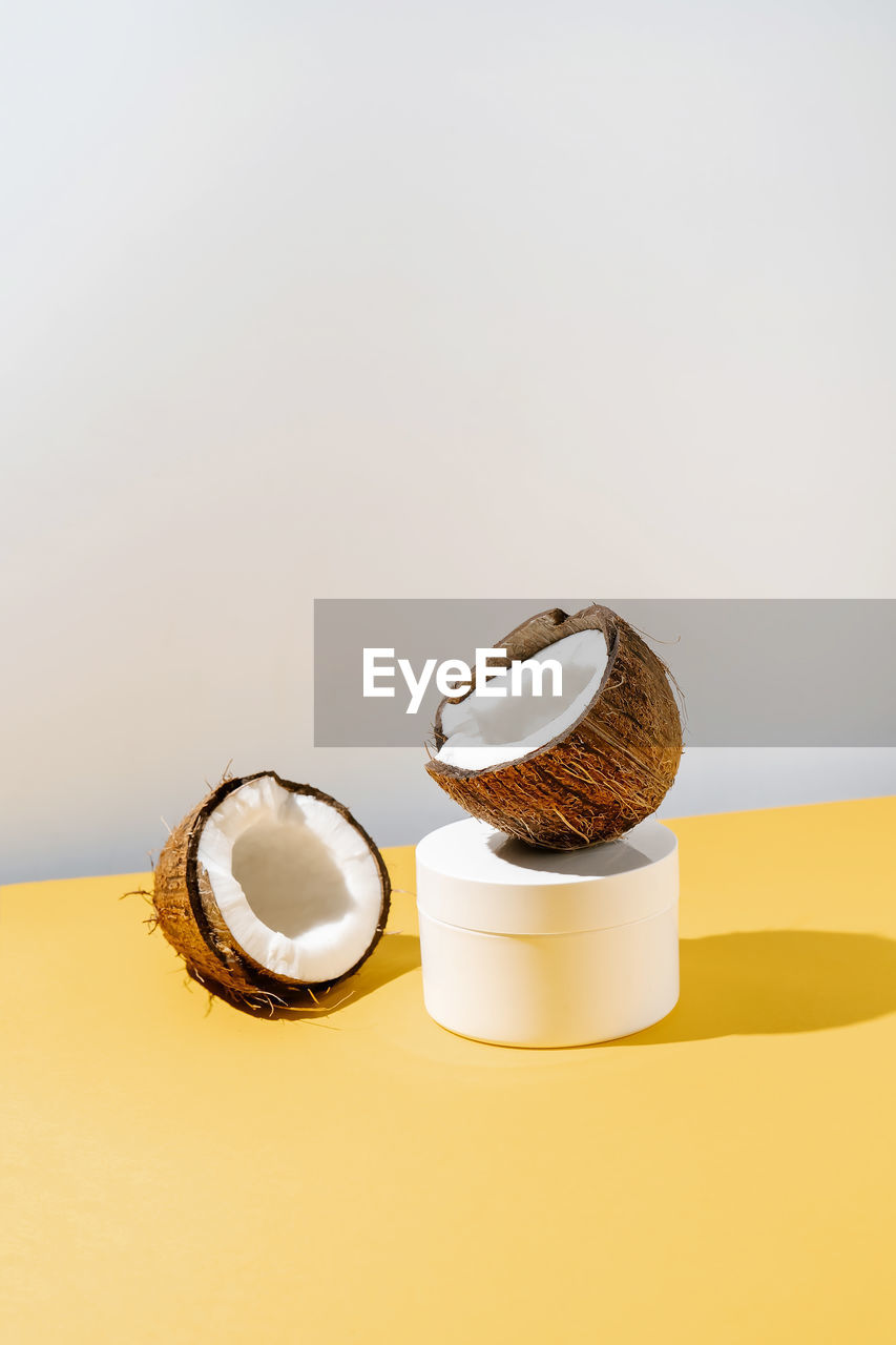 Two halves of coconut and a jar of cosmetics on a yellow and gray background.