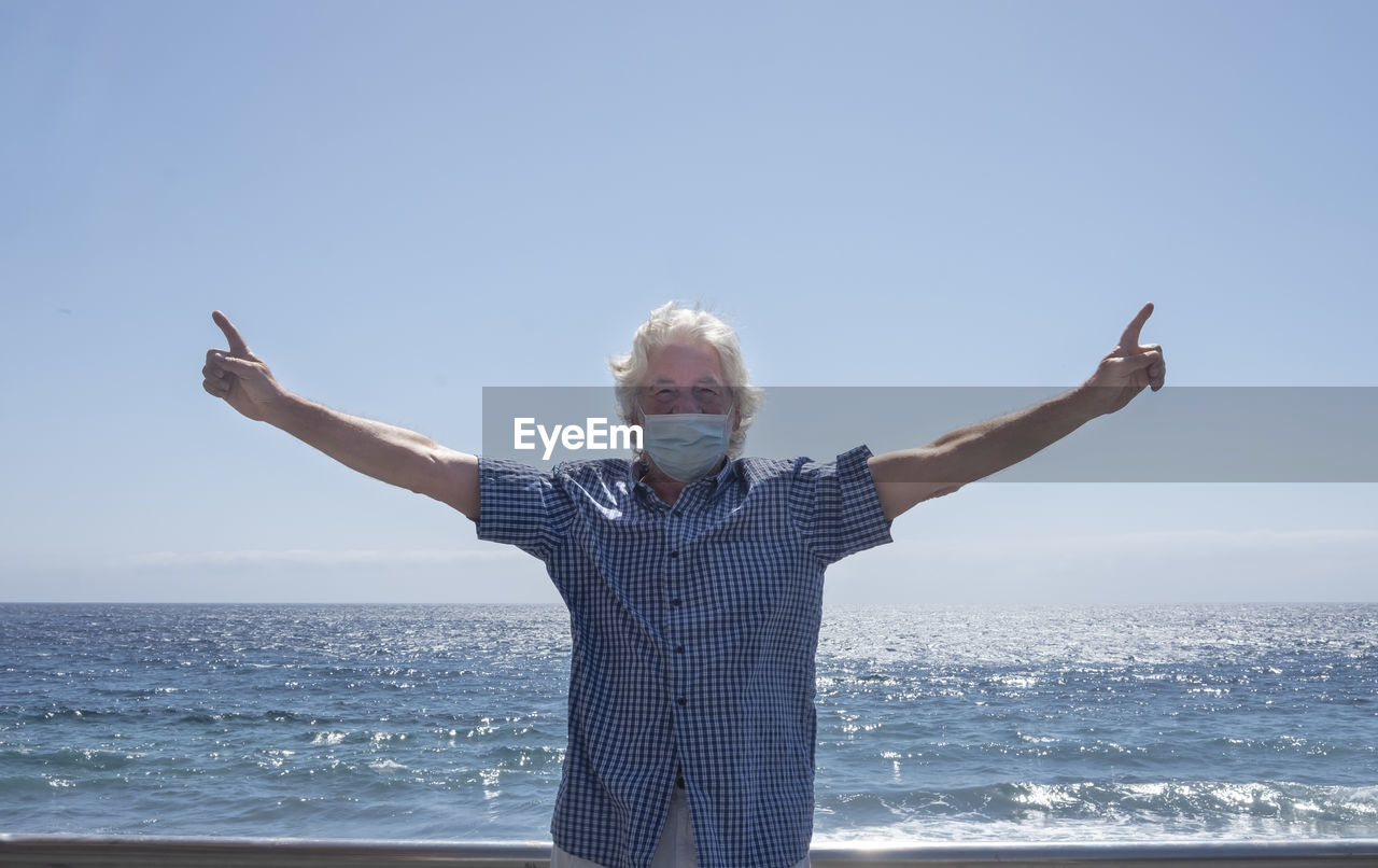 Man wearing mask with arms outstretched standing against sea and clear sky