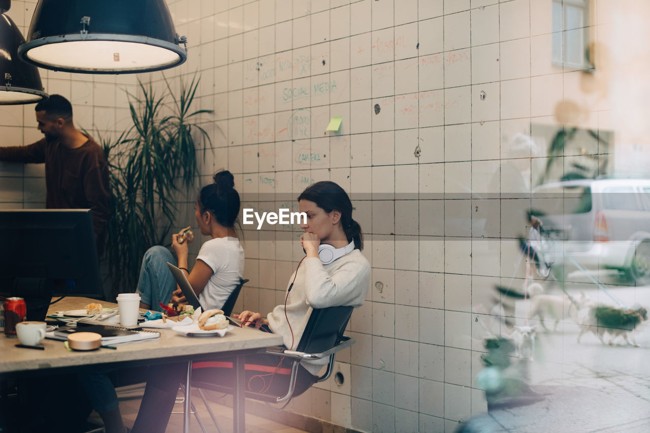 Young businesswoman using laptop while colleagues discussing at creative office seen through window