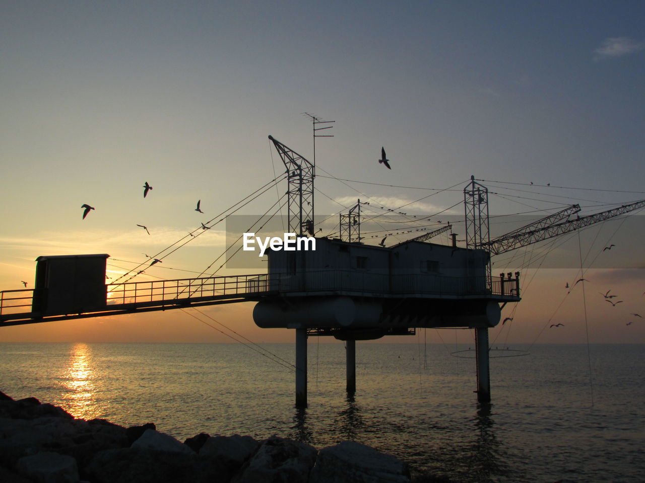 Fishing hut on stilts in sea against sky during sunset