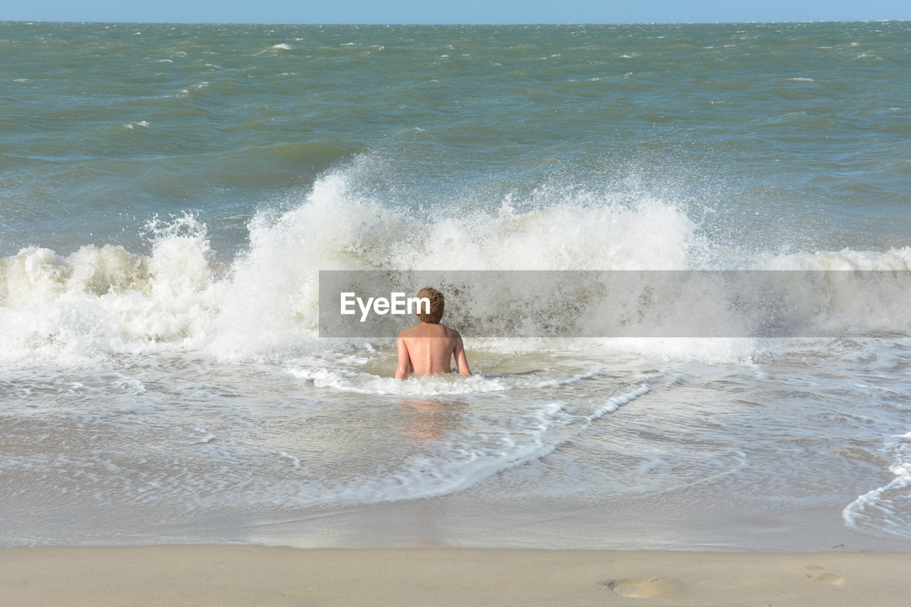 A boy sits in the water in a wave, on the sandy beach on the north sea coast in the netherlands,