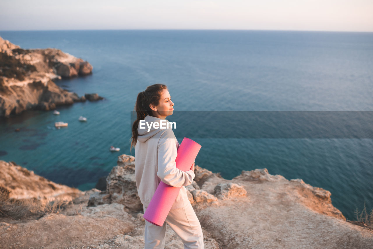 Smiling woman hold yoga mat wear sportswear hoodie and pants stand on top of rock over sea