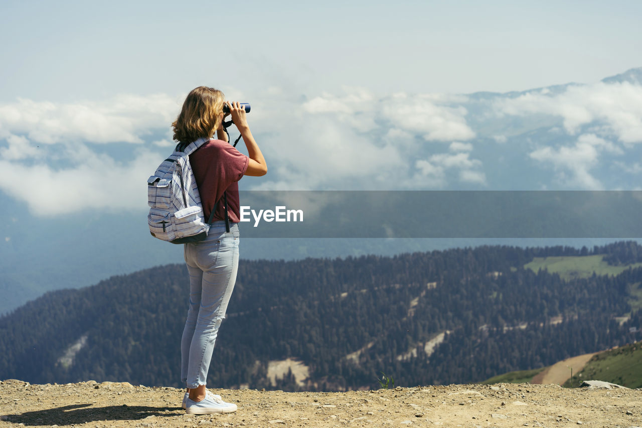 Young woman with a backpack in the mountains looks at the landscape through binoculars. 