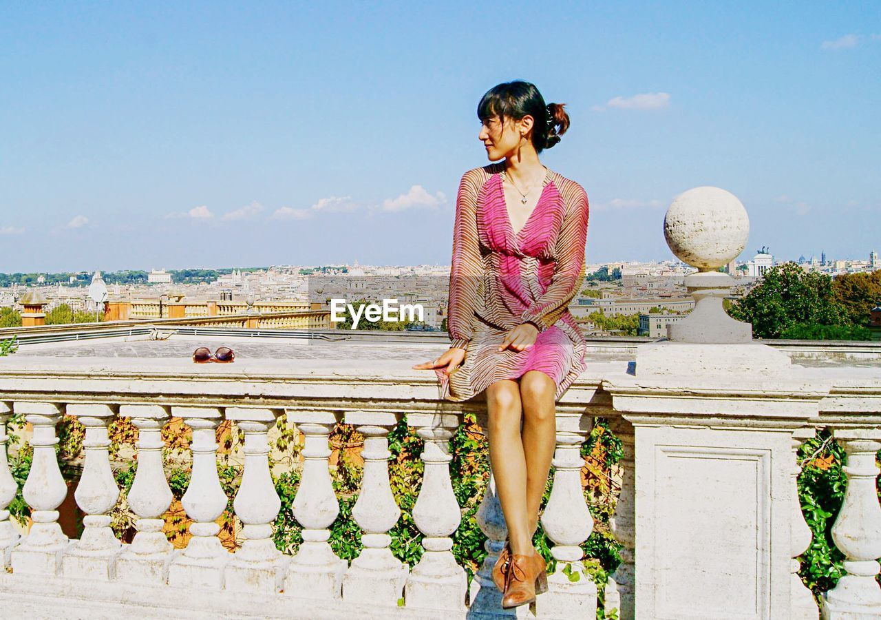 Beautiful woman sitting on balustrade against sky