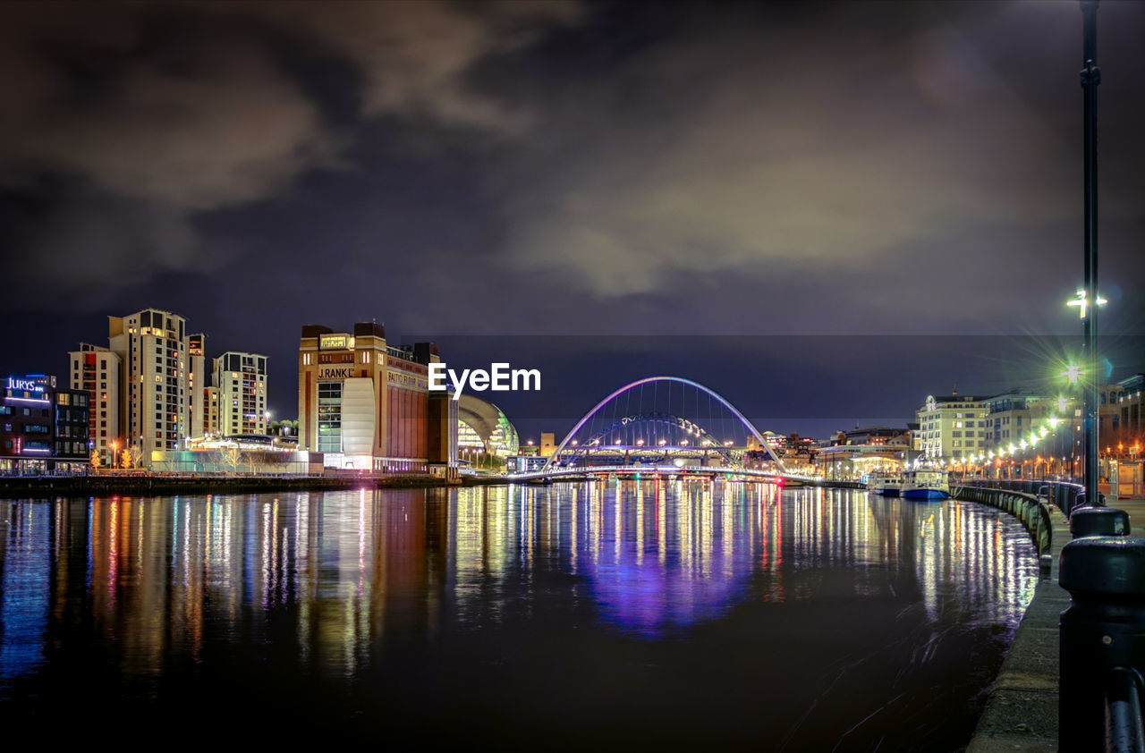 ILLUMINATED BRIDGE OVER RIVER AND BUILDINGS AGAINST SKY