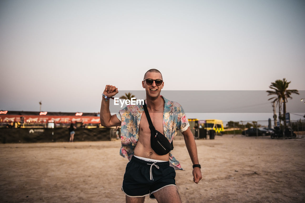 PORTRAIT OF YOUNG MAN WEARING SUNGLASSES STANDING ON BEACH