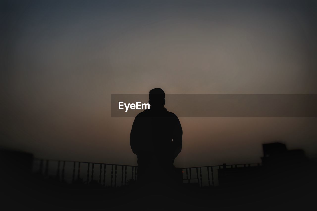 REAR VIEW OF SILHOUETTE MAN STANDING BY RAILING AGAINST SKY DURING SUNSET