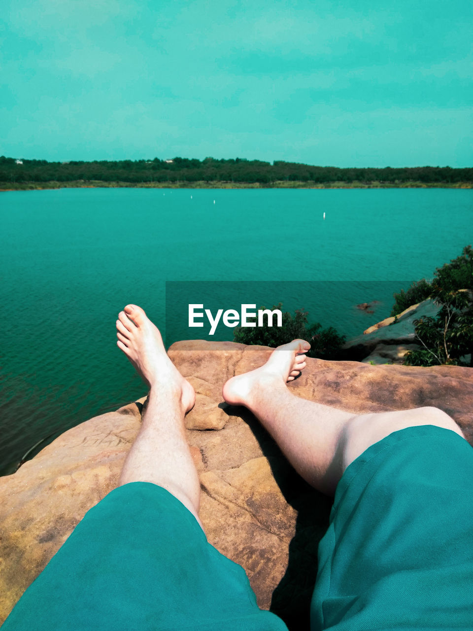 Low section of man relaxing by lake on rock during sunny day