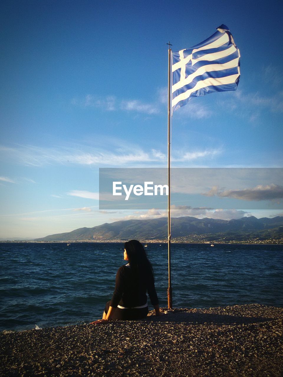 Rear view of woman sitting by greek flag on promenade against sky