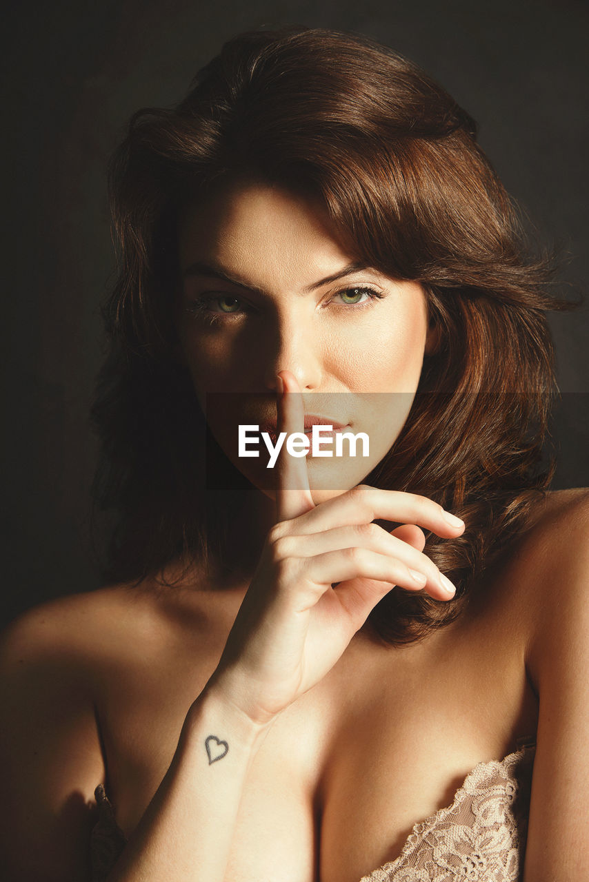 Close-up portrait of young woman with finger on lips against black background