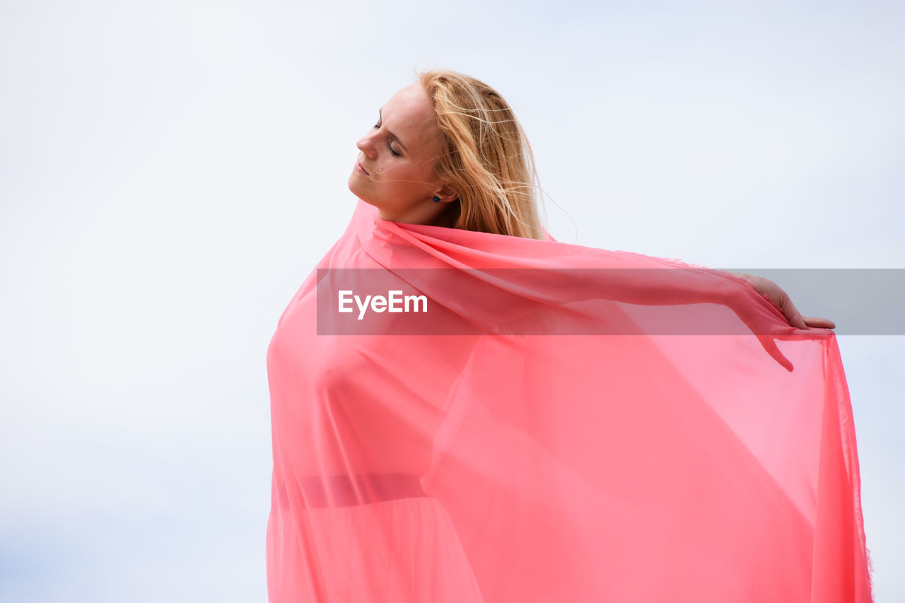 Young caucasian woman standing in desert against blue sky, covered with pink fabric.