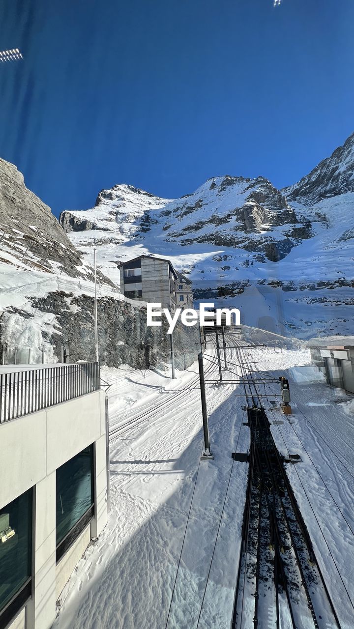 snow, cold temperature, winter, mountain, scenics - nature, nature, environment, sky, architecture, landscape, mountain range, built structure, beauty in nature, snowcapped mountain, building exterior, piste, no people, day, travel, travel destinations, building, outdoors, winter sports, frozen, resort, sunlight, tranquility, blue, tranquil scene, land, tourism, house, water