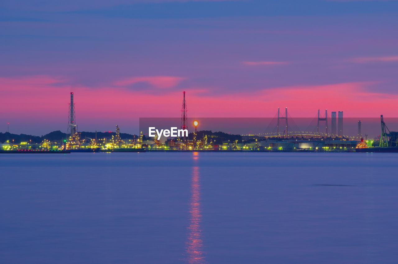 Illuminated city by sea against sky at sunset