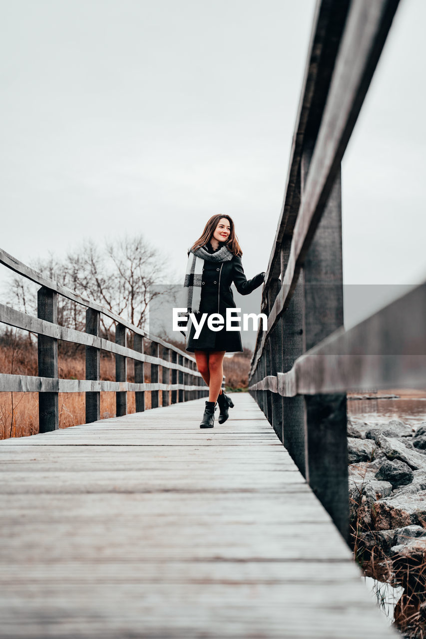 WOMAN STANDING ON FOOTBRIDGE AGAINST SKY