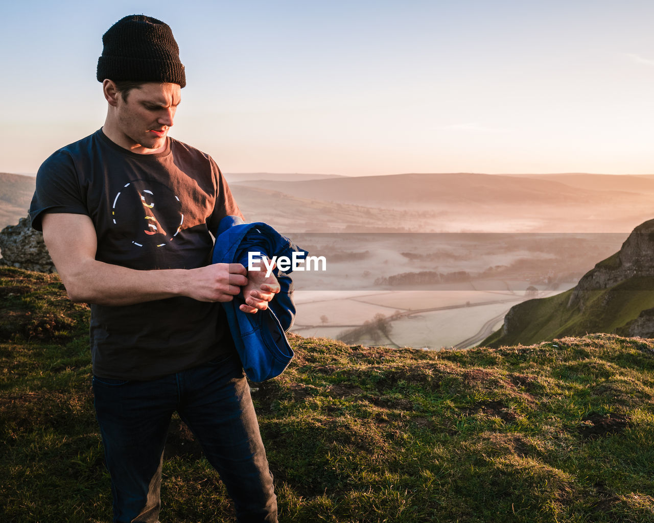 Man holding jacket while standing on mountain against sky during sunset