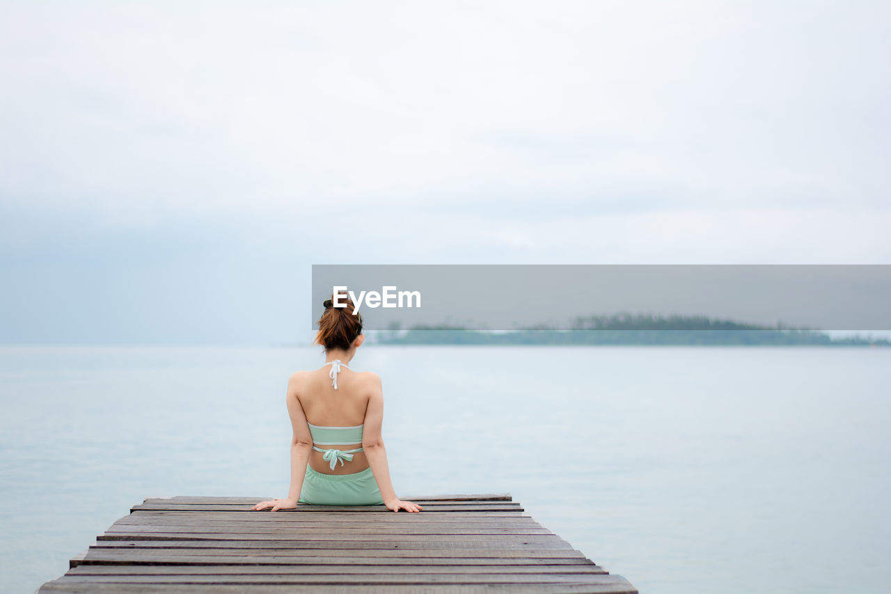 Rear view of woman sitting on pier over sea against sky