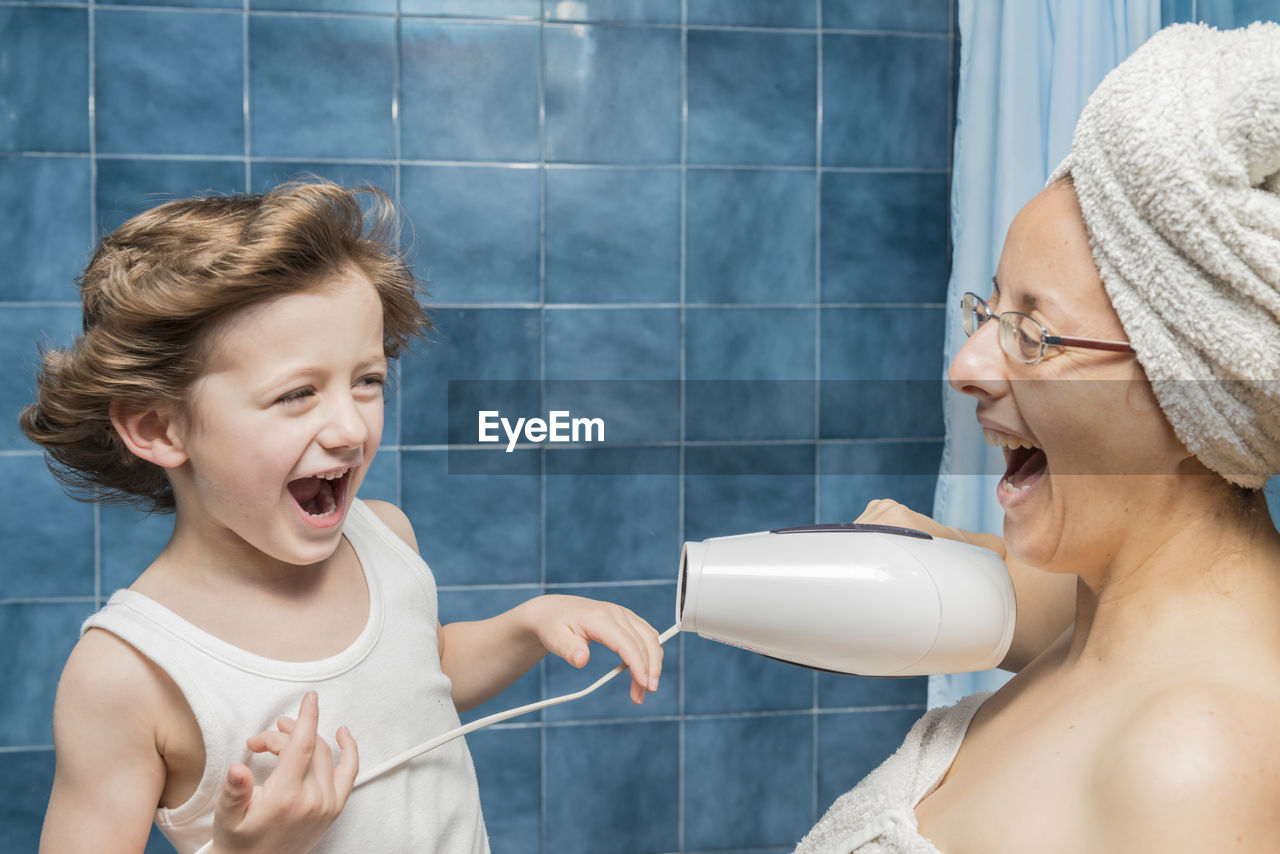 Cheerful mother blowing air with dryer on son face in bathroom