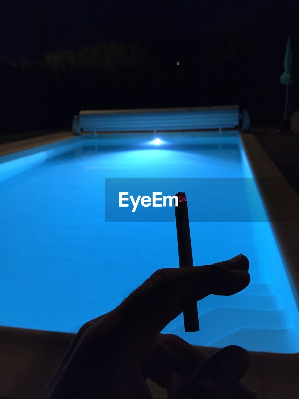 Cropped image of person holding cigarette against swimming pool