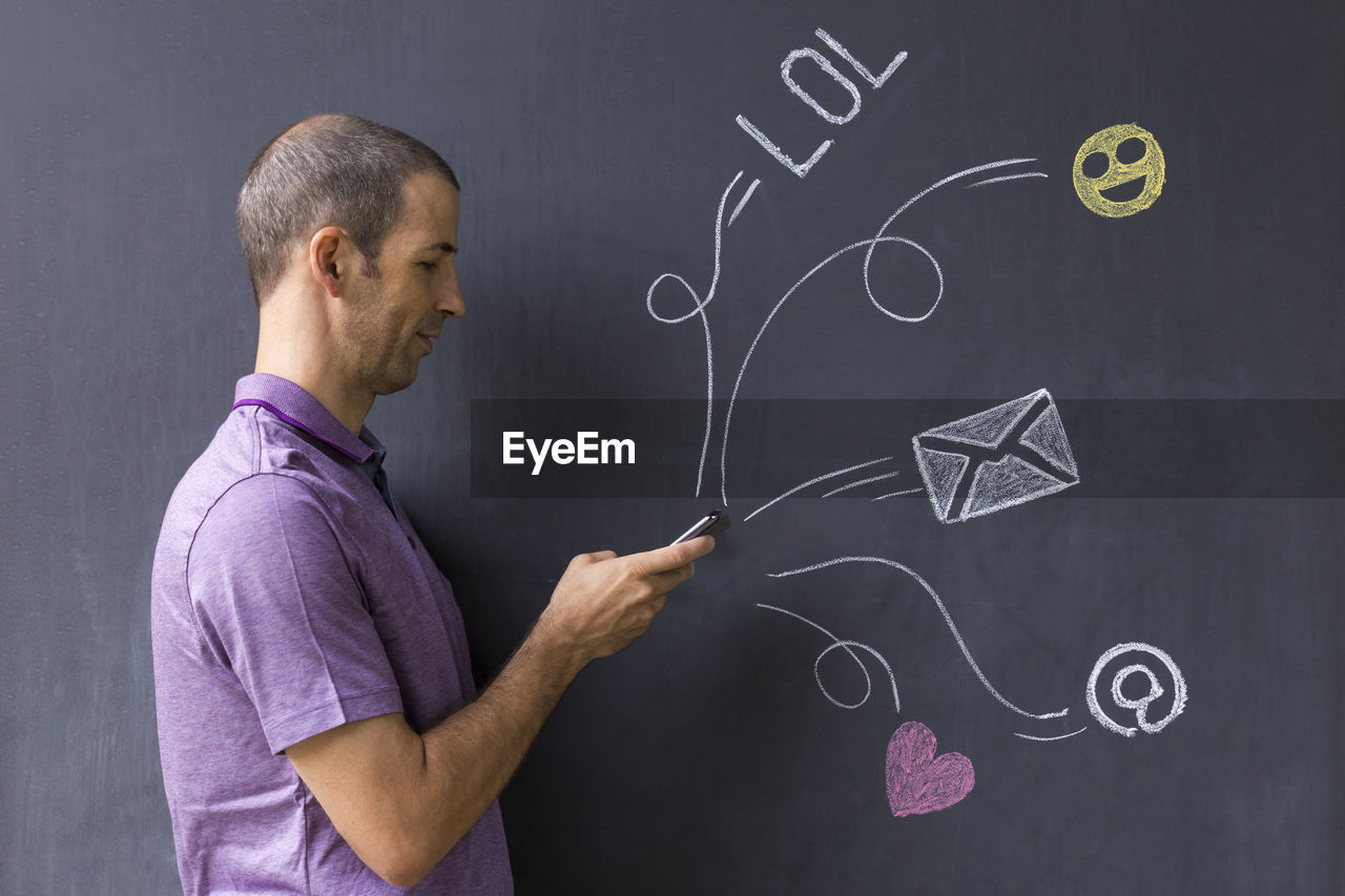 Side view man using mobile phone while standing by blackboard with various icons