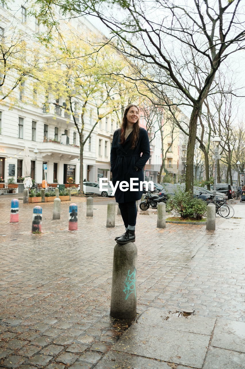 Full length of young woman standing on bollard over walkway in city