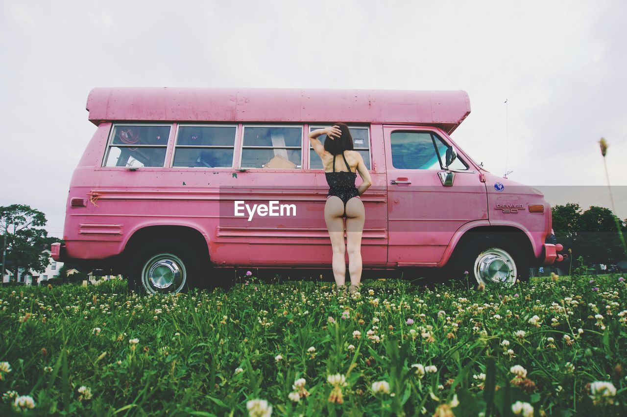 Rear view of young woman in bikini standing by car on grassy field against clear sky
