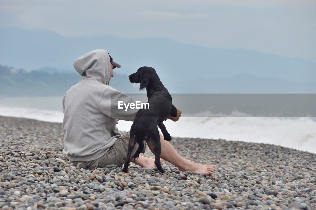 Man with dog sitting at beach against sky
