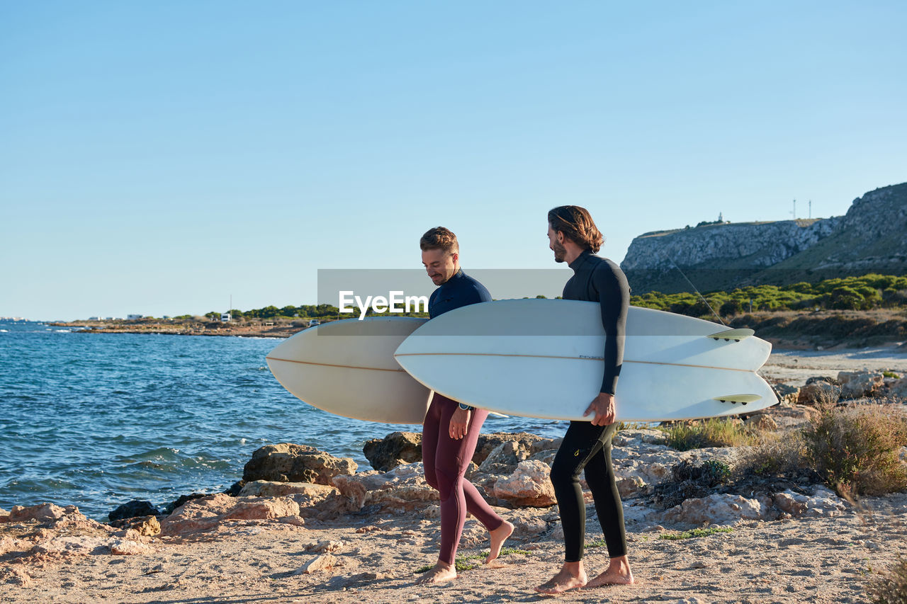 Positive male surfers in wetsuits and with surfboards walking along wet seashore after surfing at sunset