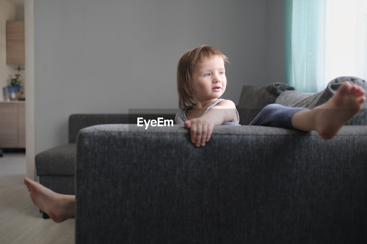 European toddler child sits and gets bored on gray sofa in bright living room, minimalist interior