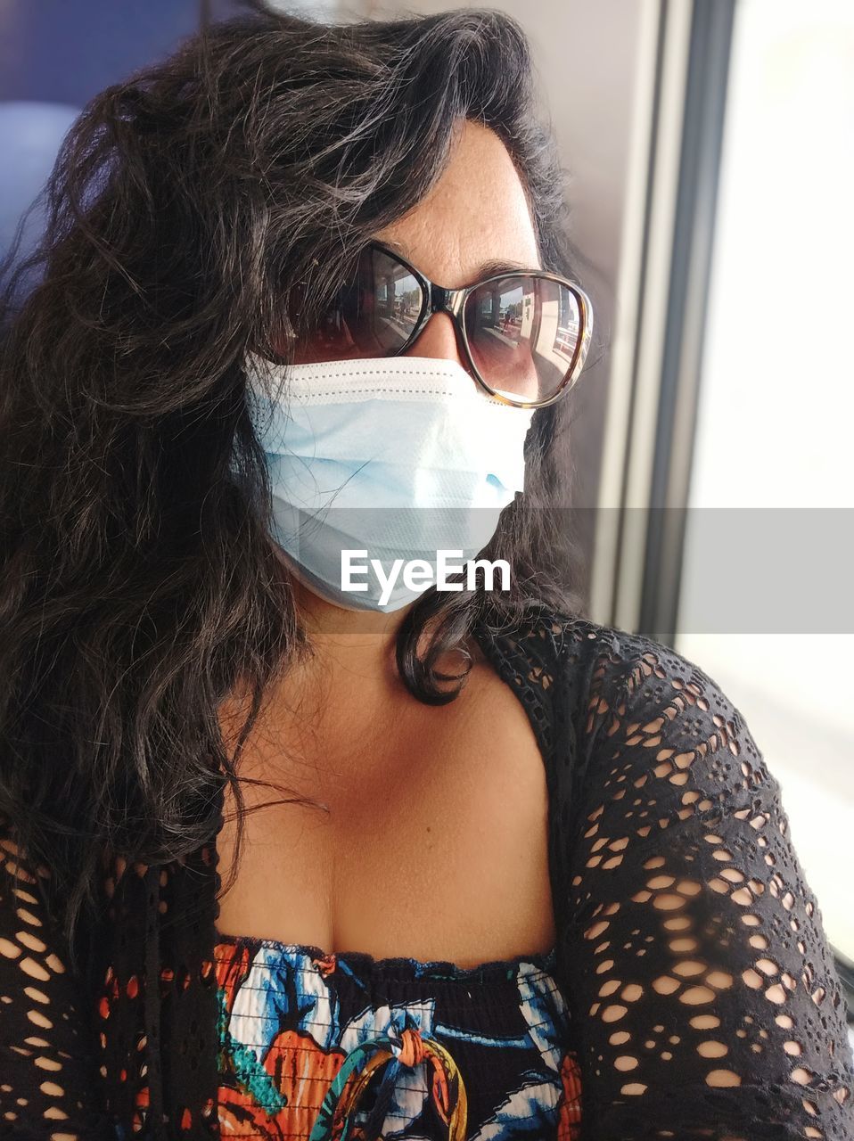 Portrait of carefree woman wearing sunglasses and protective face mask while sitting by window