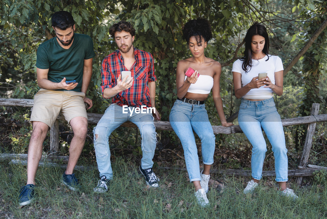 Full length of friends using smart phones while sitting on railing against trees