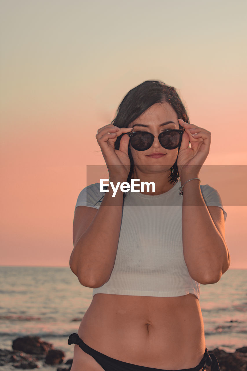 Portrait of woman wearing sunglasses while standing at beach against sky during sunset